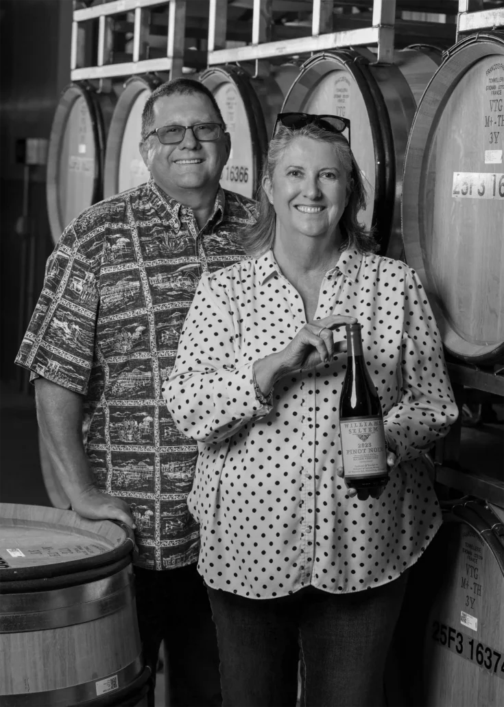 John & Diane Bucher stand in front of wine barrels; the woman holds a bottle of wine and both are smiling at the camera.