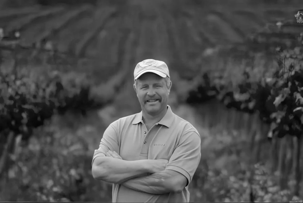 A man wearing a cap and polo shirt stands with arms crossed in a vineyard, with grapevines and rows of crops in the background.