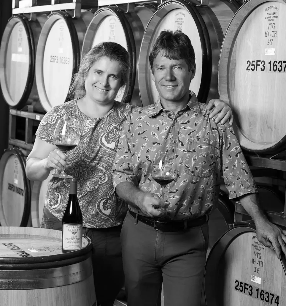 The Schoeneman Family stand in front of wine barrels, each holding a glass of wine. A wine bottle is placed on a barrel in front of them.
