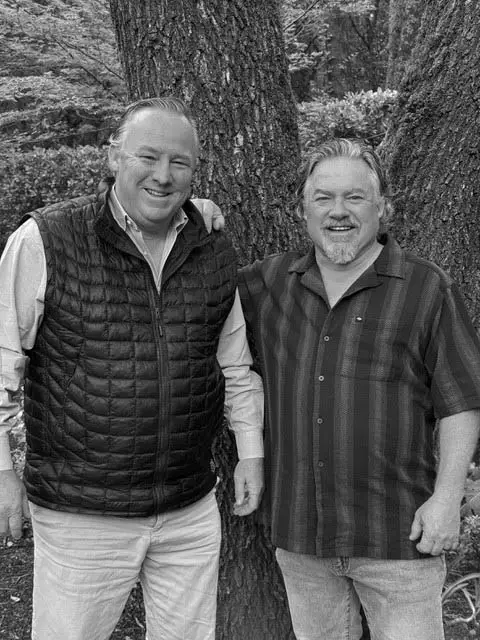 Donnie and Bix Weir stand next to each other outdoors in front of a tree, smiling at the camera. One wears a quilted vest, the other a short-sleeve striped shirt. The photo is in black and white.