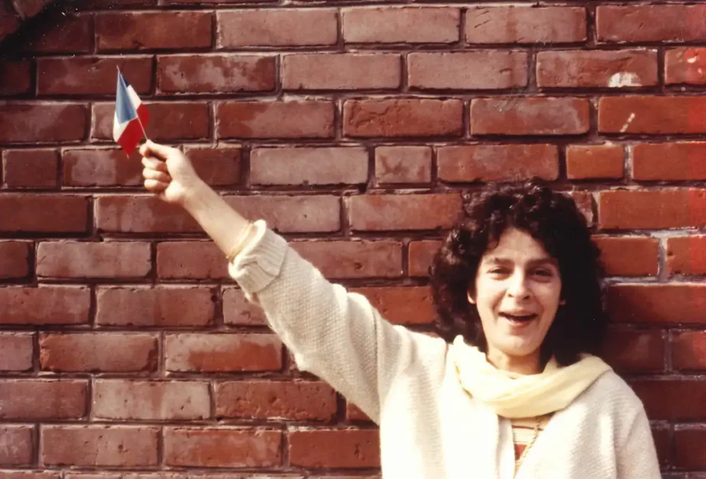 Becky Wasserman stands in front of a brick wall, smiling and holding up a small French flag.