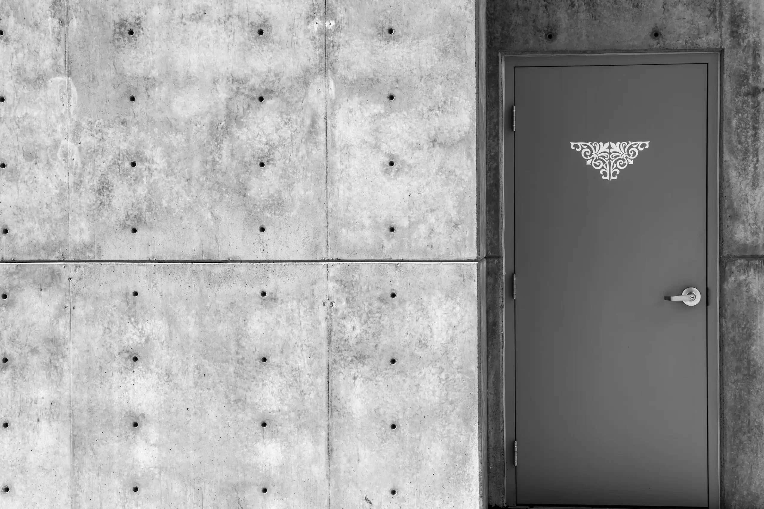 A plain gray door with a decorative white pattern at the top, set in a concrete wall with visible bolts and seams.