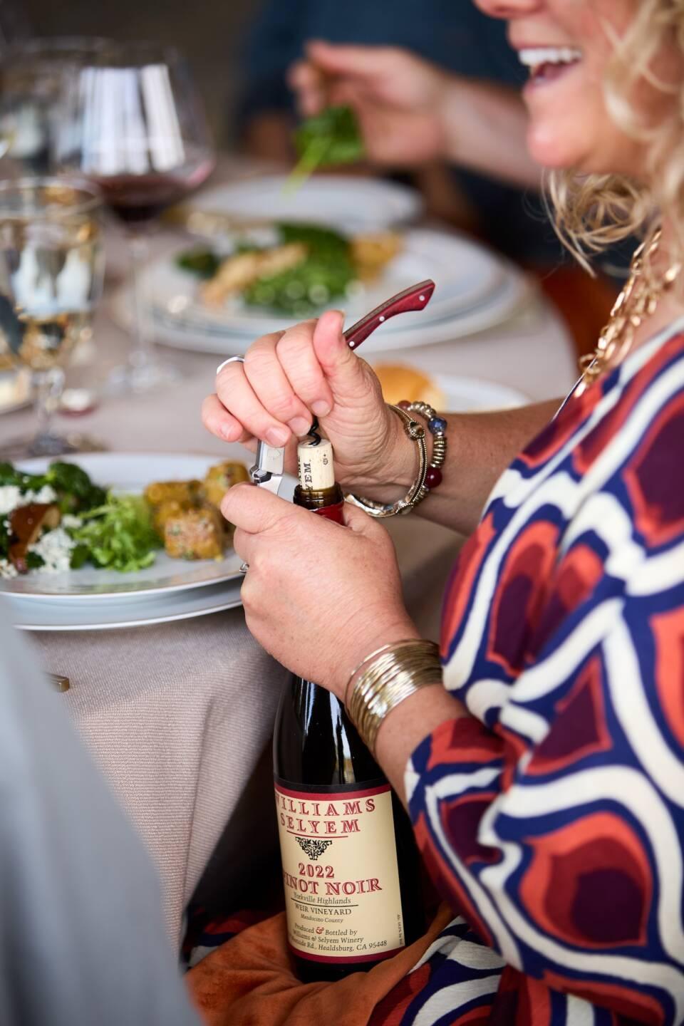 A woman in a patterned dress opens a bottle of 2022 Williams Selyem Pinot Noir at a dining table set with plates of food and glasses of wine.