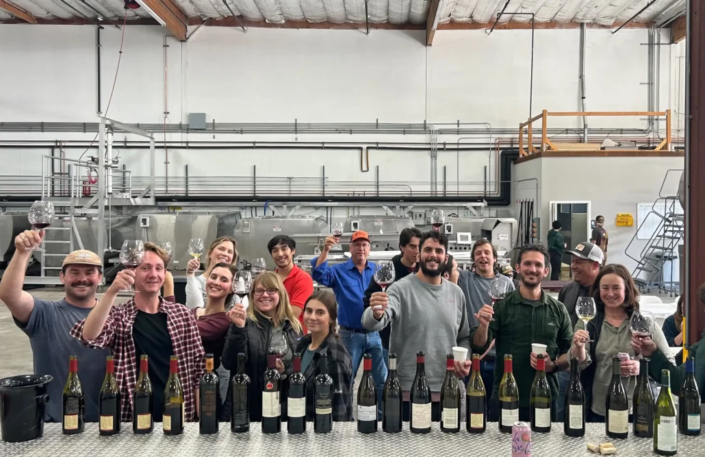 A group of people are holding up wine glasses and bottles, smiling in a winery or production facility.