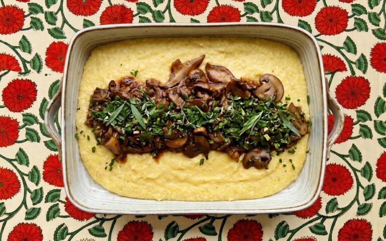 A rectangular dish of creamy polenta topped with sautéed mushrooms and chopped herbs, set on a table with a red floral patterned tablecloth.