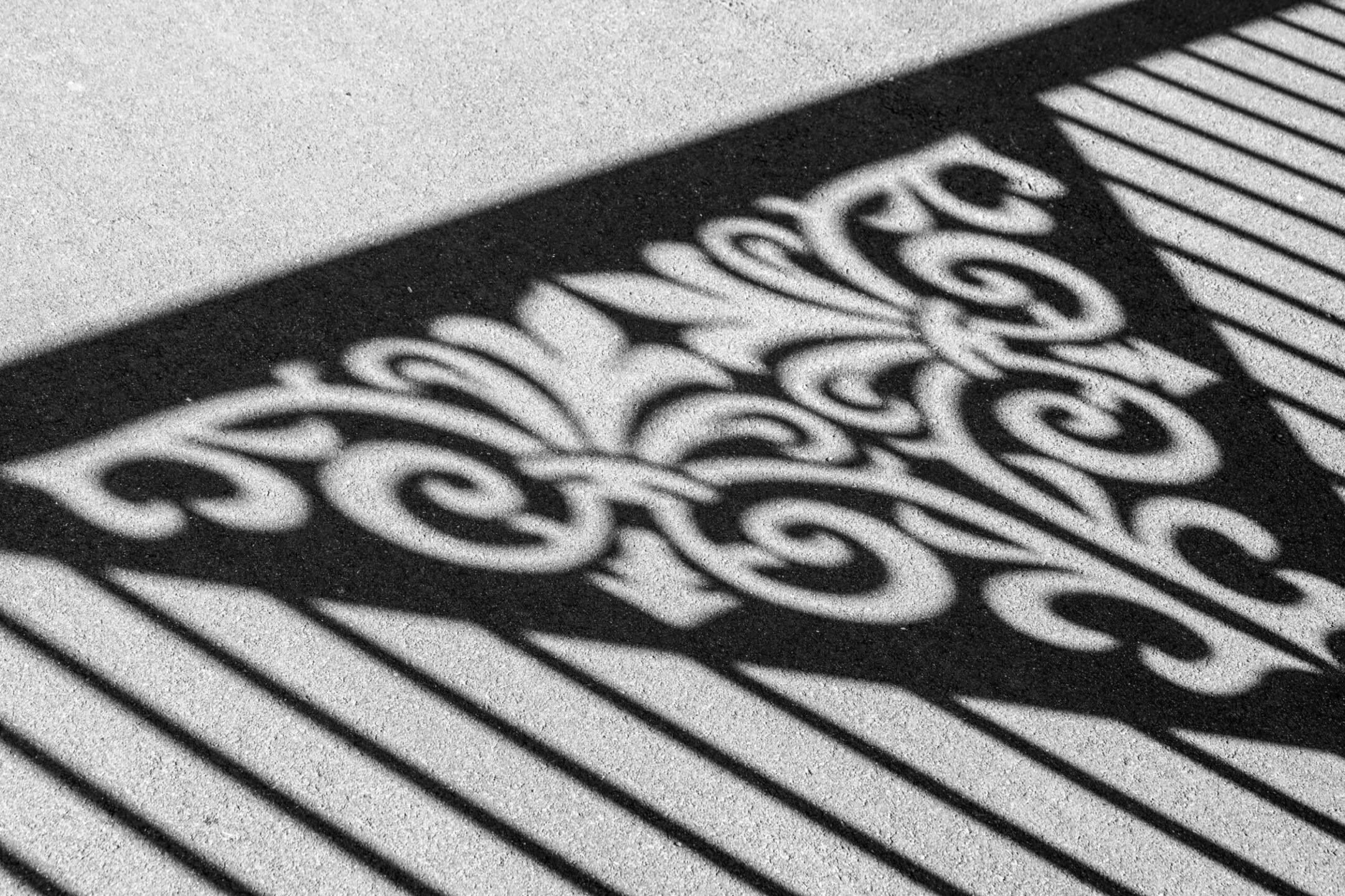 Shadow of an ornate metal fence with decorative scrollwork cast onto a light-colored concrete surface.