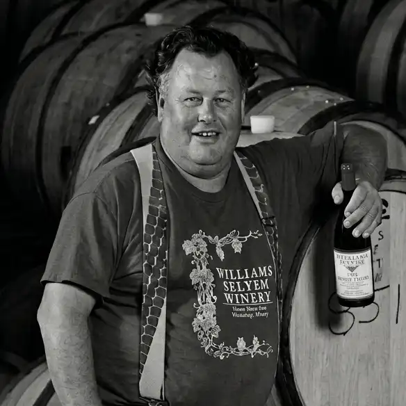 Founder Burt Williams stands in front of wine barrels, holding a bottle of wine and smiling at the camera.
