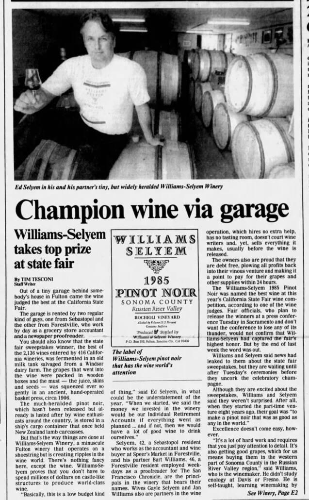 A man sits on a table in a wine cellar with wooden barrels behind him; newspaper article discusses Williams-Selyem Winery's award-winning 1985 Pinot Noir.