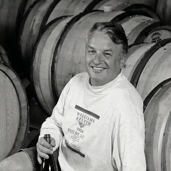Founder Ed Selyem holding a wine bottle sits in front of stacked wooden barrels in a winery.