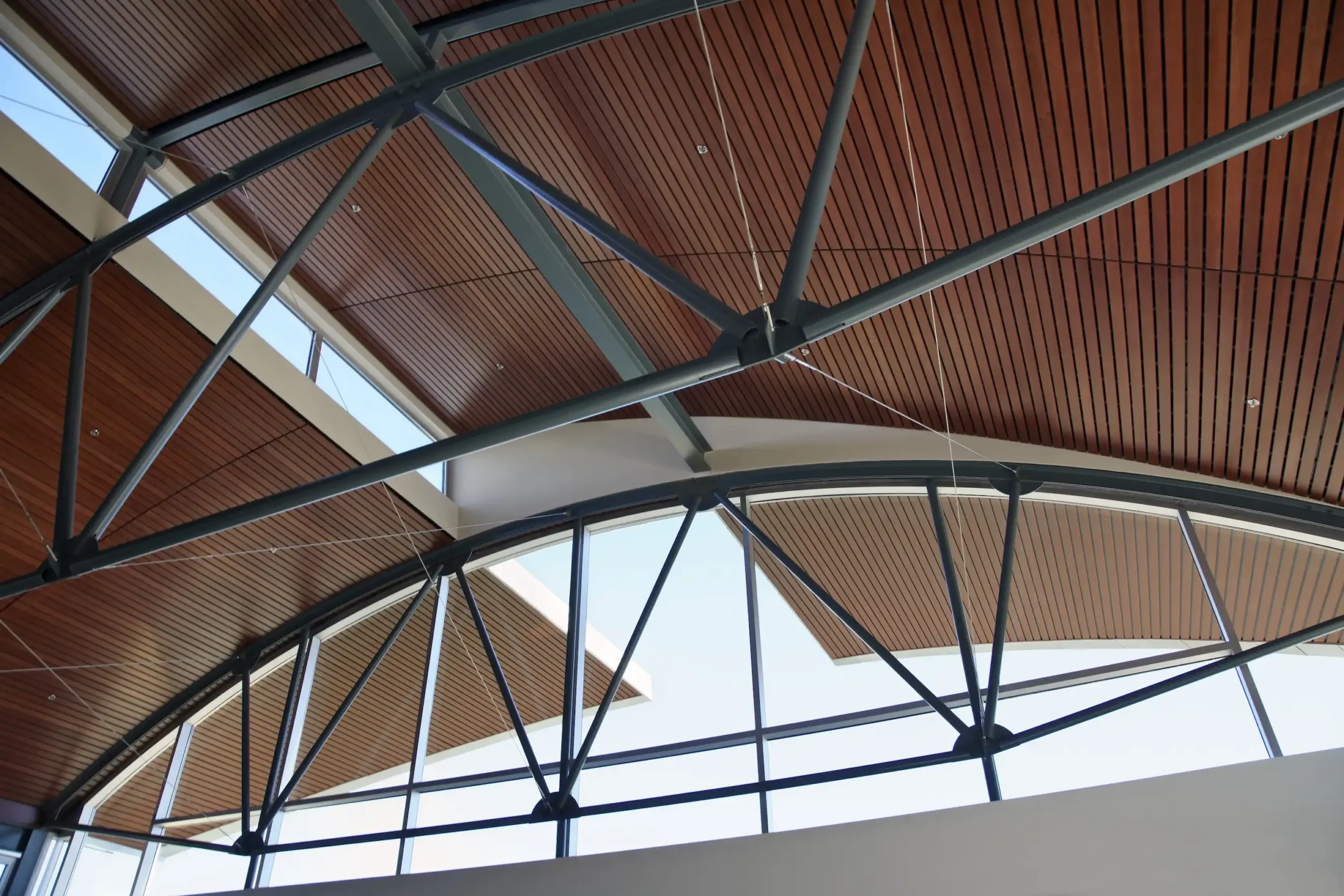 Interior view of a modern building's ceiling with exposed steel beams, wooden panels, and large arched windows letting in natural light.