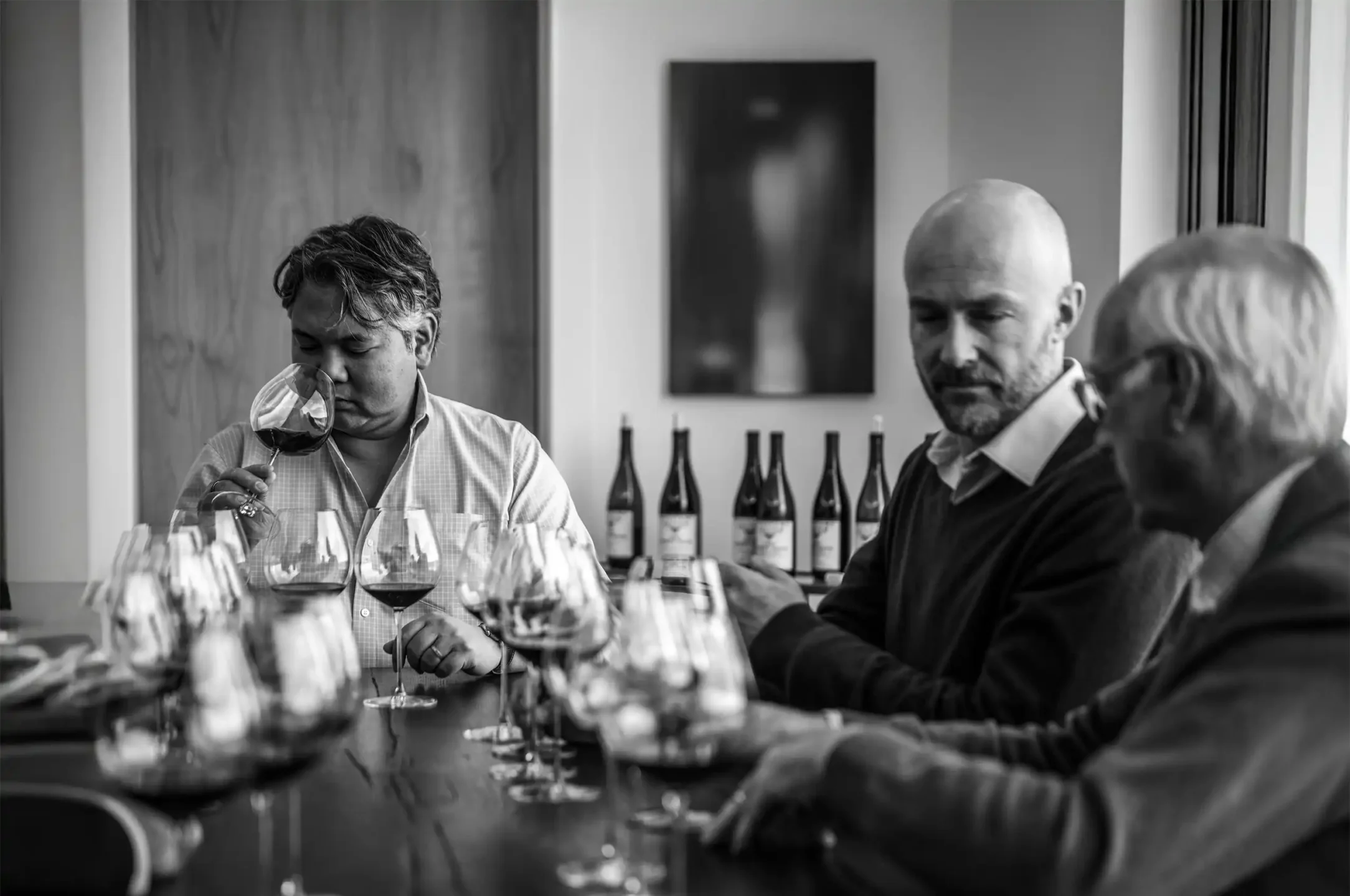 Jeff Mangahas, Erwan Faiveley, and John Dyson sit at a table with several wine glasses, one man smelling wine, in a room with bottles lined up in the background.