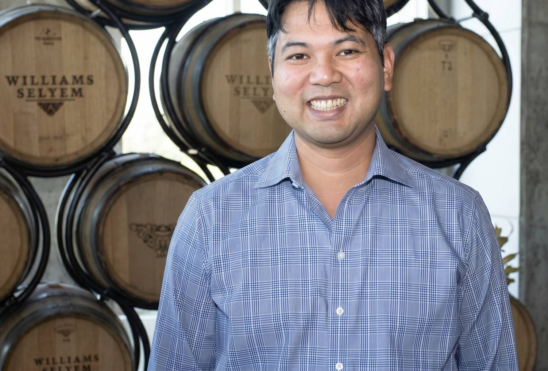 Jeff Mangahas in a blue checked shirt stands smiling in front of stacked wine barrels labeled "Williams Selyem.