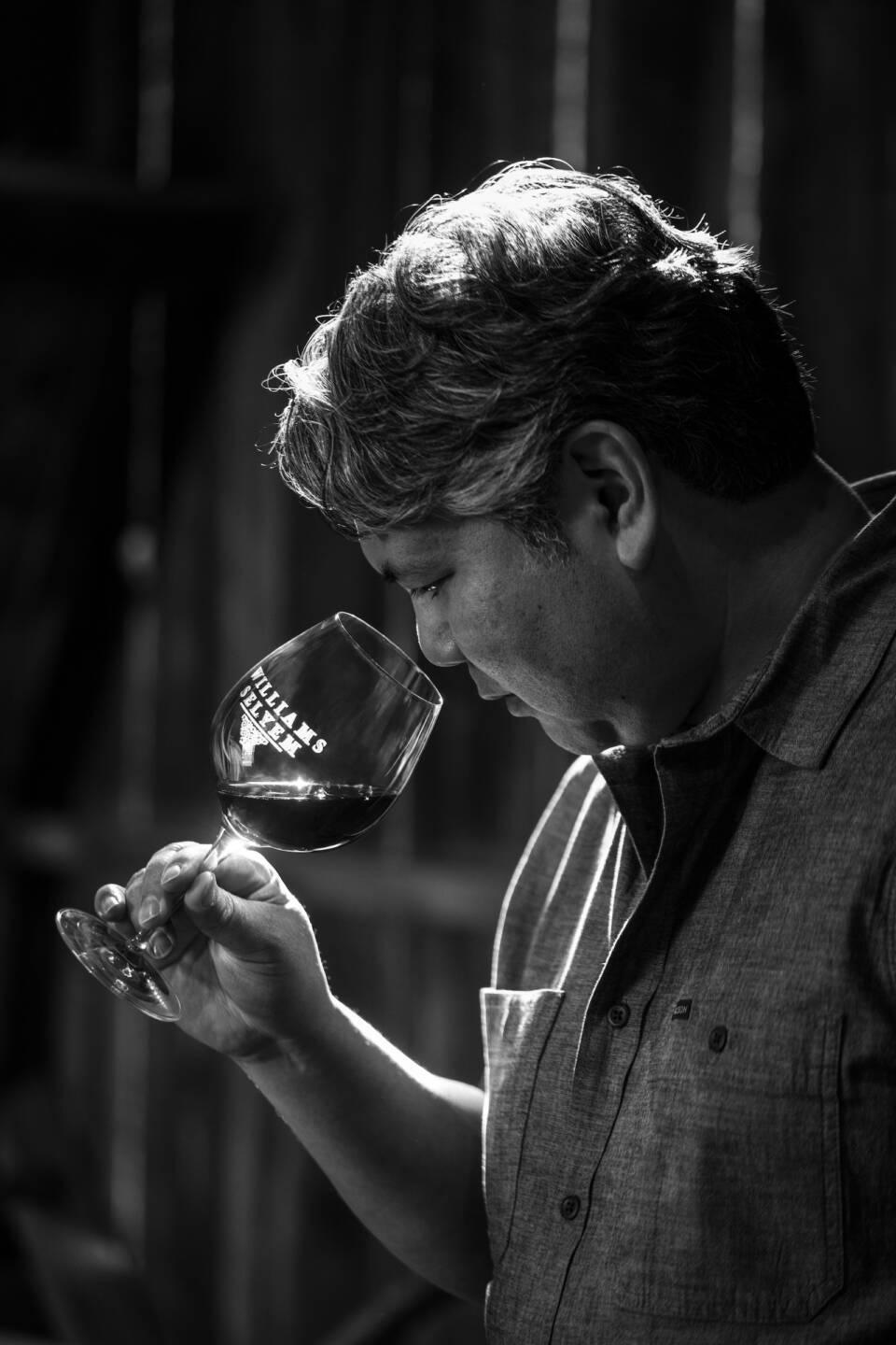 Williams Selyem Vice President and Director of Winemaking Jeff Mangahas holds a wine glass up to his nose and appears to be smelling the wine in the cellar. The image is black and white.