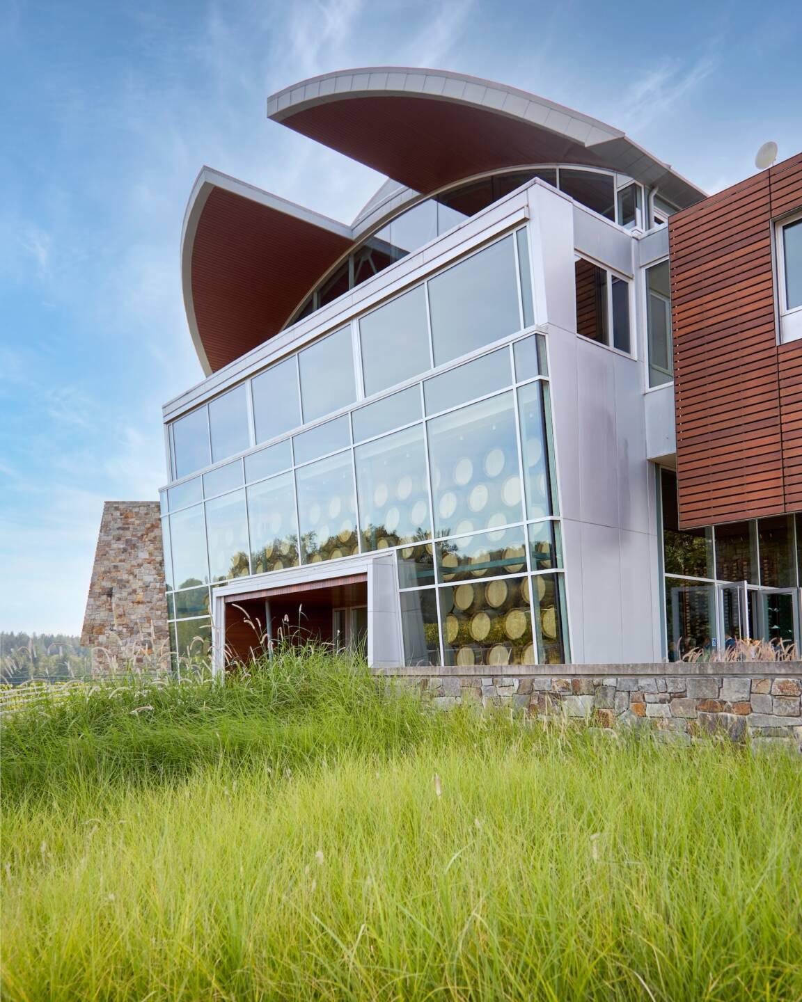 Modern building with large glass windows, distinctive curved roof, and stone accent wall, set behind tall green grass under a blue sky.