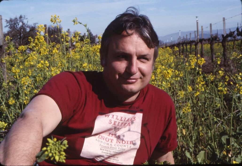 A man wearing a red T-shirt sits in a field of yellow flowers with grapevines and mountains in the background under a clear sky.