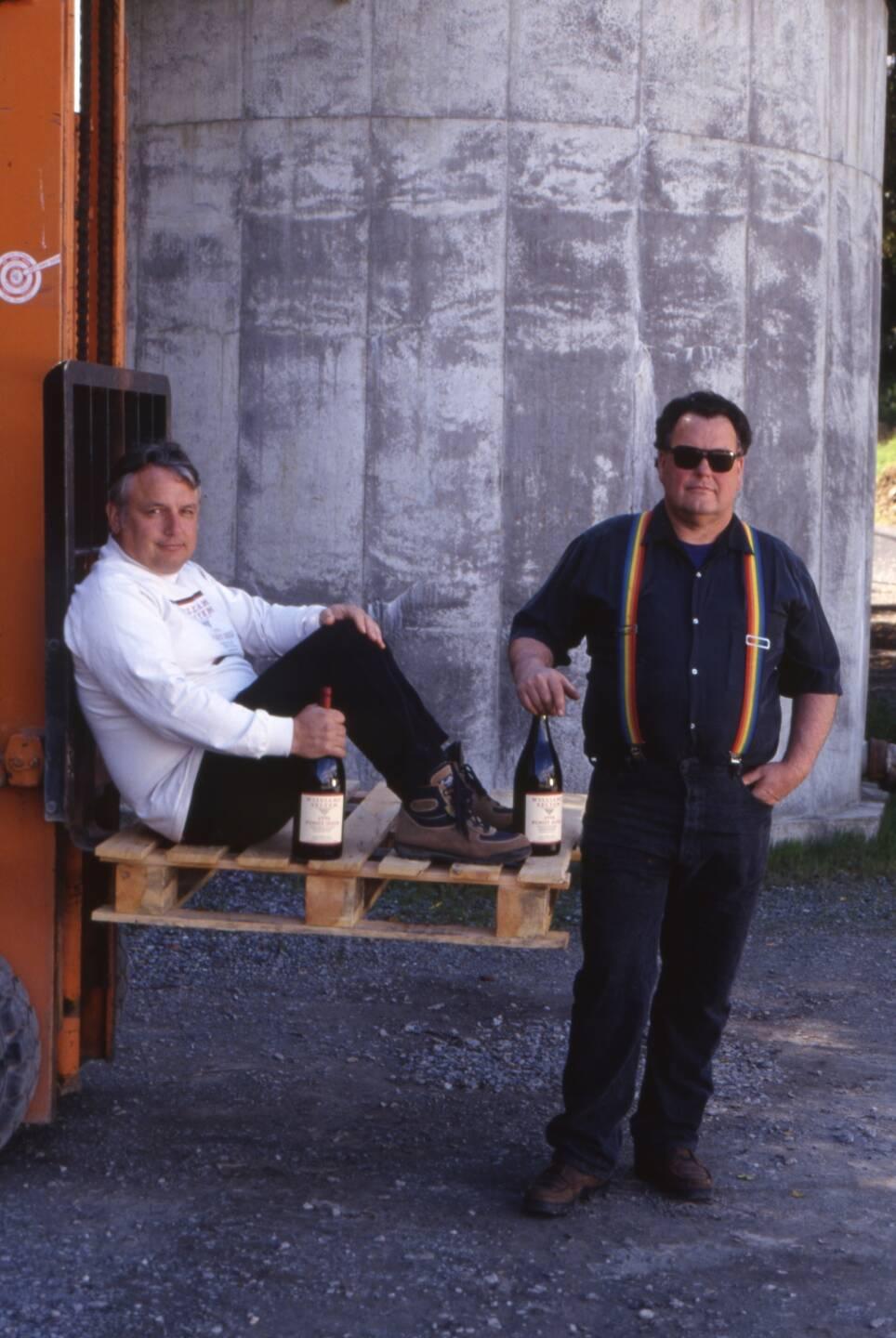 Two men pose with wine bottles; one sits on a forklift pallet, the other stands beside him. They are outdoors in front of a large concrete structure.