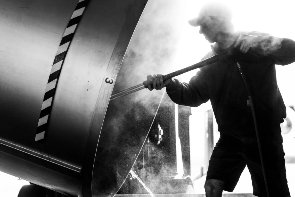 A person in a cap stirs or shovels inside a large, steaming metal container marked with the number 3, with strong backlighting and visible steam.