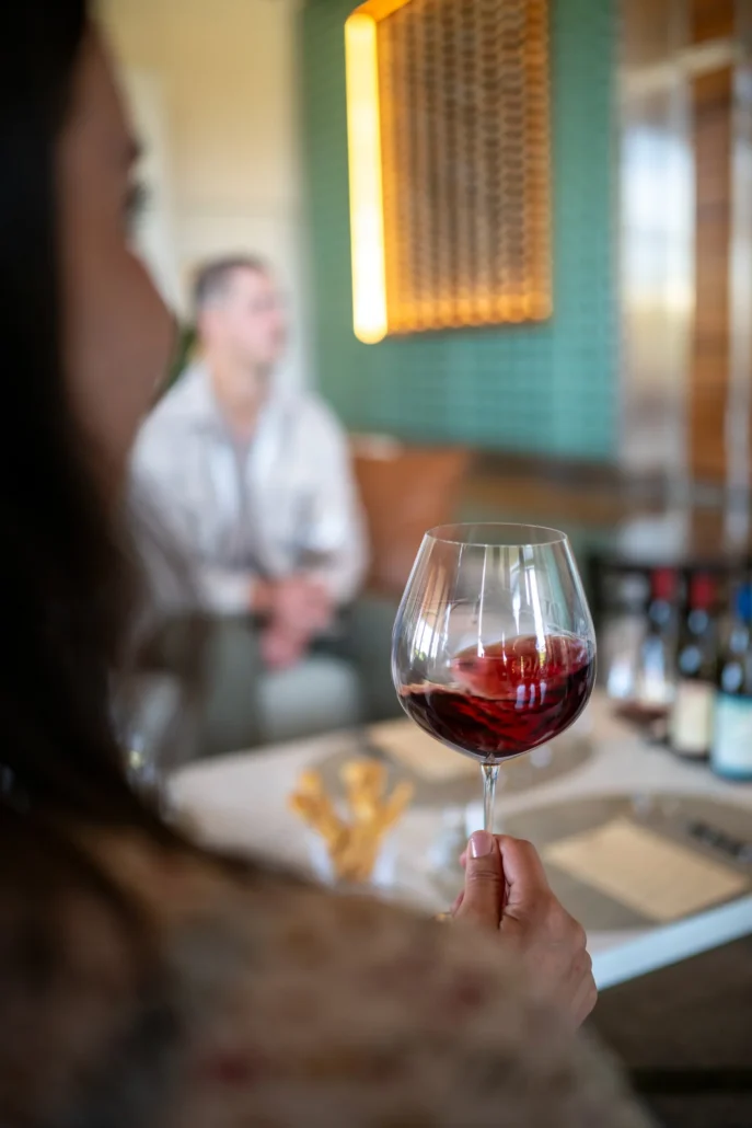 A person holds a glass of red wine in the foreground, with another person and several wine bottles blurred in the background.