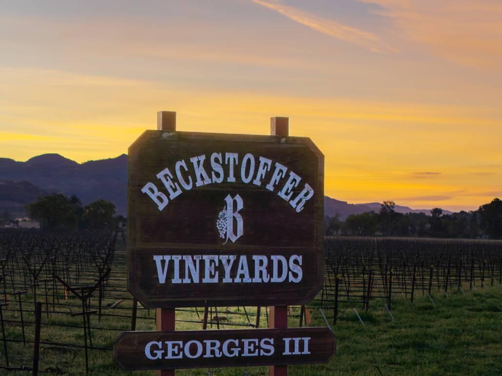 A wooden sign reading "Beckstoffer Vineyards Georges III" stands in front of a vineyard at sunset, with mountains in the background.