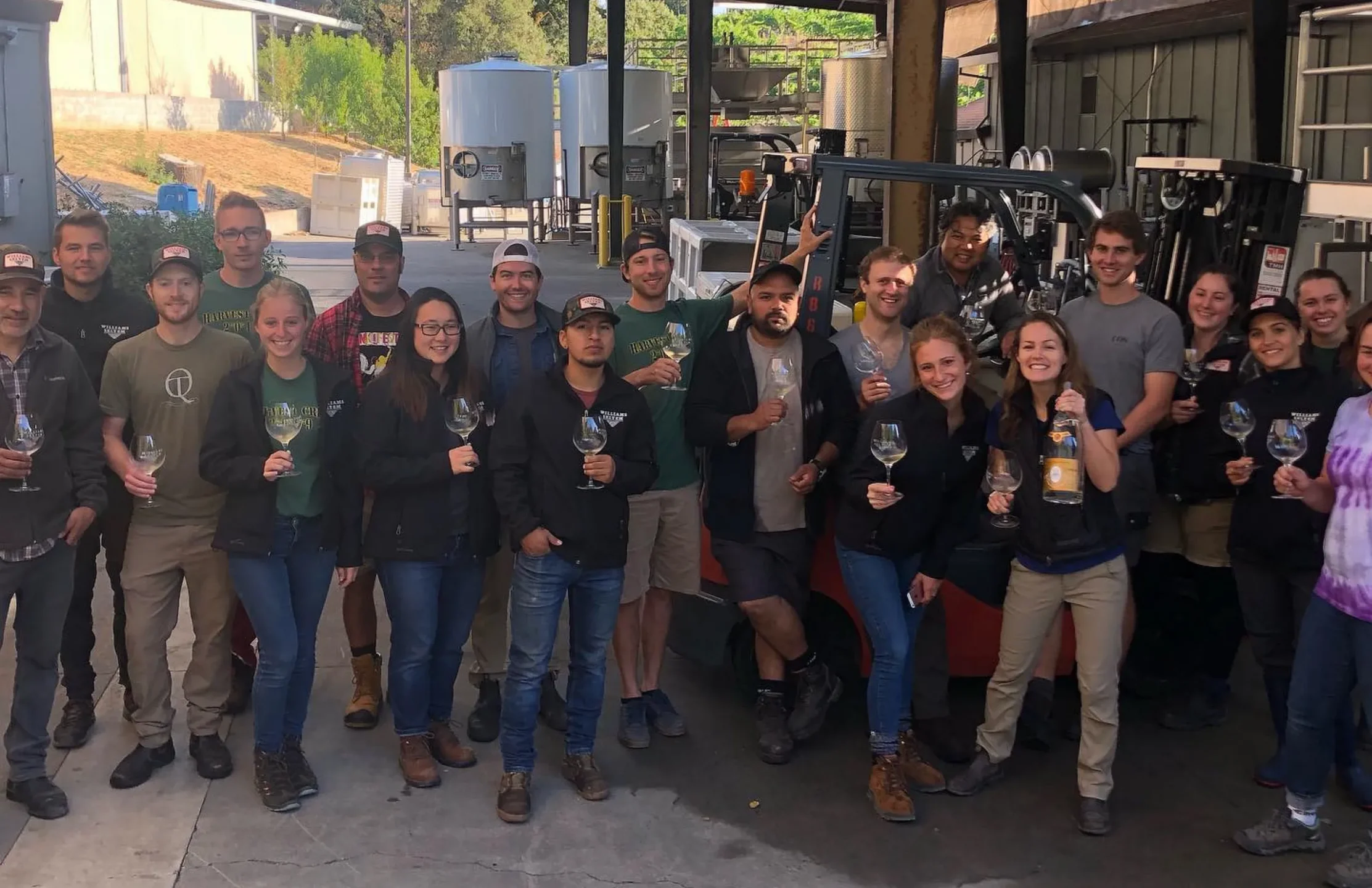 A group of people, many holding wine glasses, pose together outdoors in an industrial setting with equipment and buildings in the background.