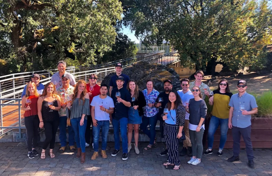 A group of 20 people stand outdoors on a stone patio, smiling for a group photo; most are holding drinks and trees are in the background.