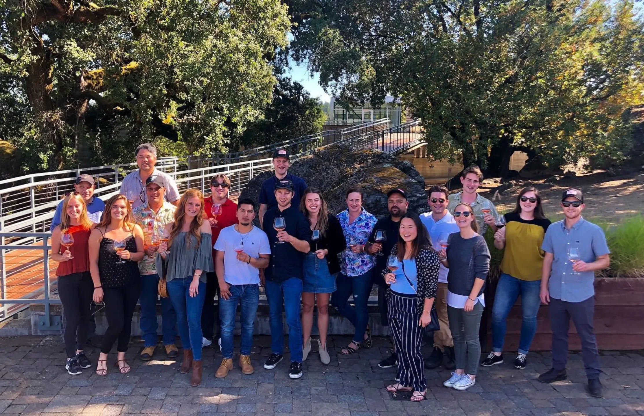 A group of 20 people stand outdoors on a stone patio, smiling for a group photo; most are holding drinks and trees are in the background.