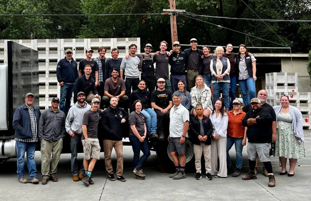 A large group of people poses together outdoors in front of stacked crates and a truck, smiling at the camera. Trees and utility lines are visible in the background.