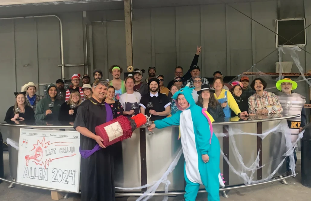 A group of people in costumes and casual wear pose behind a decorated bar with a "Last Grill 2024" sign; two individuals stand in front, one in a dinosaur suit and one holding a large bottle prop.