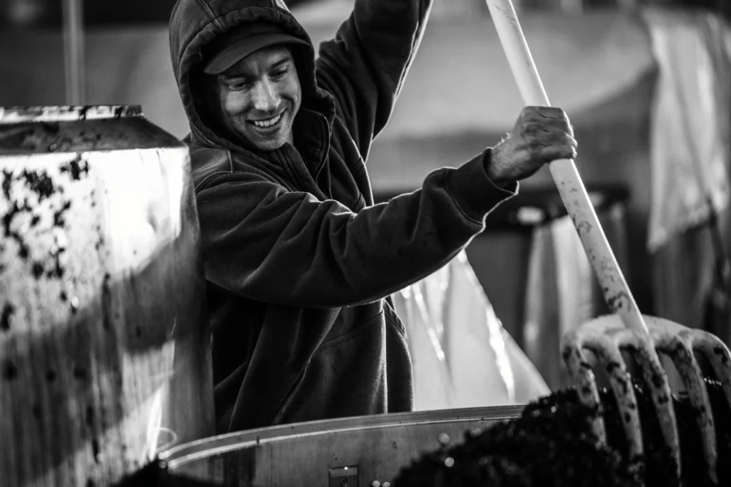 A person in a hoodie uses a large tool to stir or move grapes in a container, smiling while working in what appears to be a winery or production facility.