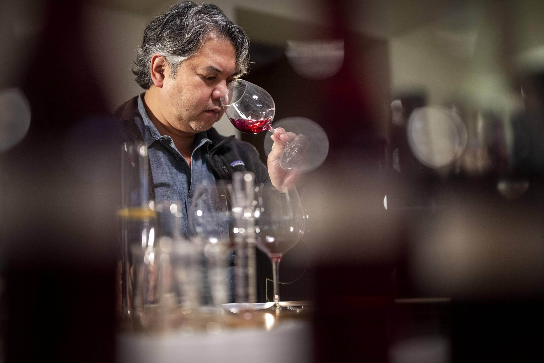 Williams Selyem Vice President and Director of Winemaking Jeff Mangahas smells a glass of red wine, surrounded by blurred bottles and glasses on a table.