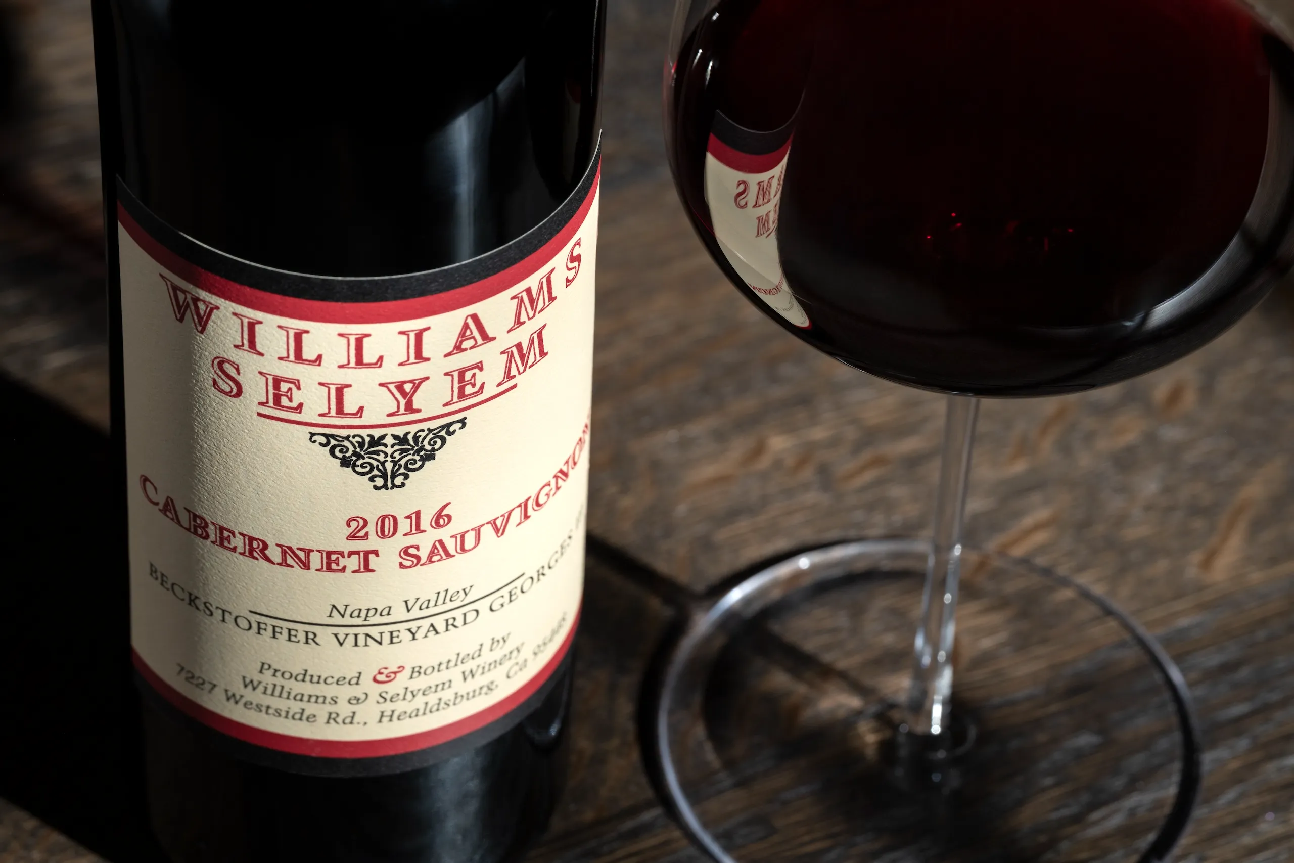 A bottle of 2016 Williams Selyem Cabernet Sauvignon and a glass of red wine on a wooden surface.