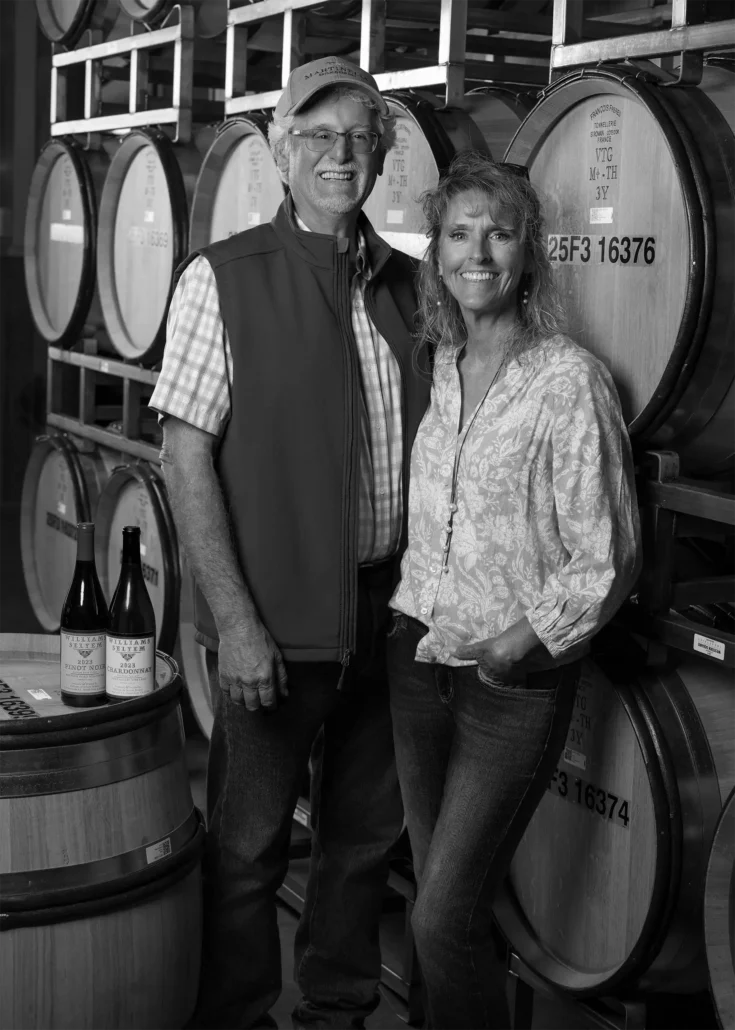 A man and woman stand together in a winery, smiling in front of stacked wine barrels. Two wine bottles and glasses rest on a nearby barrel.