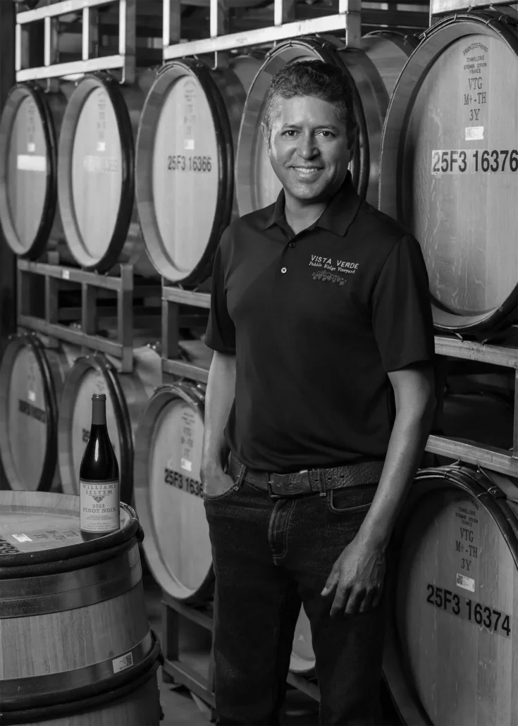 Miguel Alvarado in a Vista Verde Vineyards polo shirt stands in a wine cellar beside stacked wine barrels, with a wine bottle and glass on a barrel in front of him.