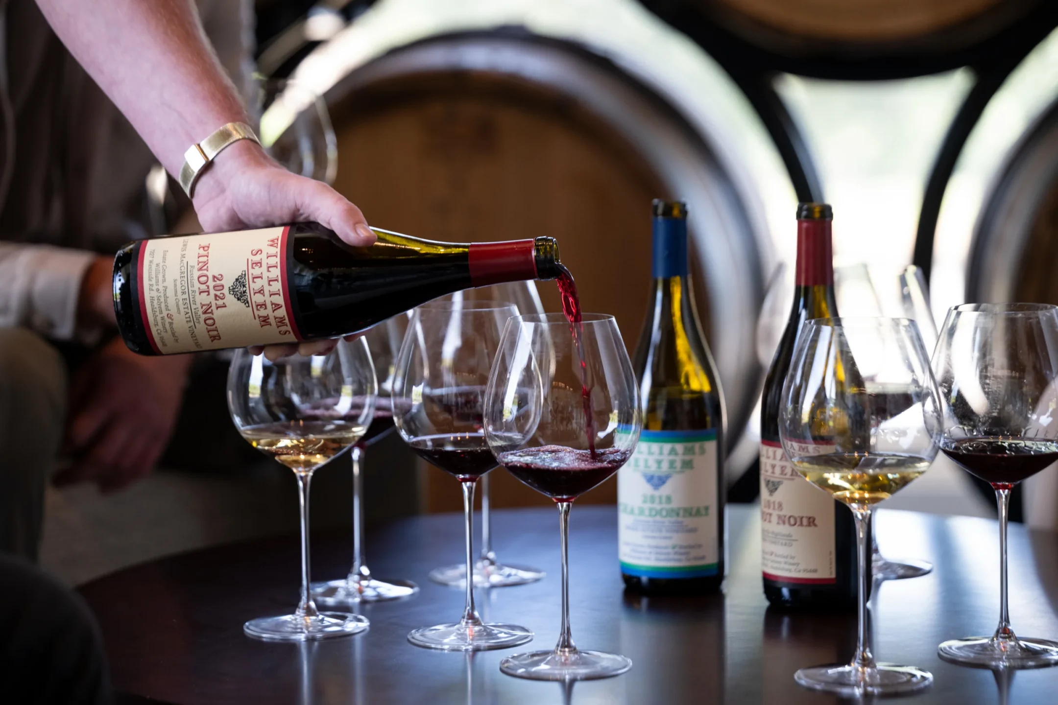 A hand pours red wine from a bottle into a glass on a table with several wine glasses and bottles, with wine barrels in the background.