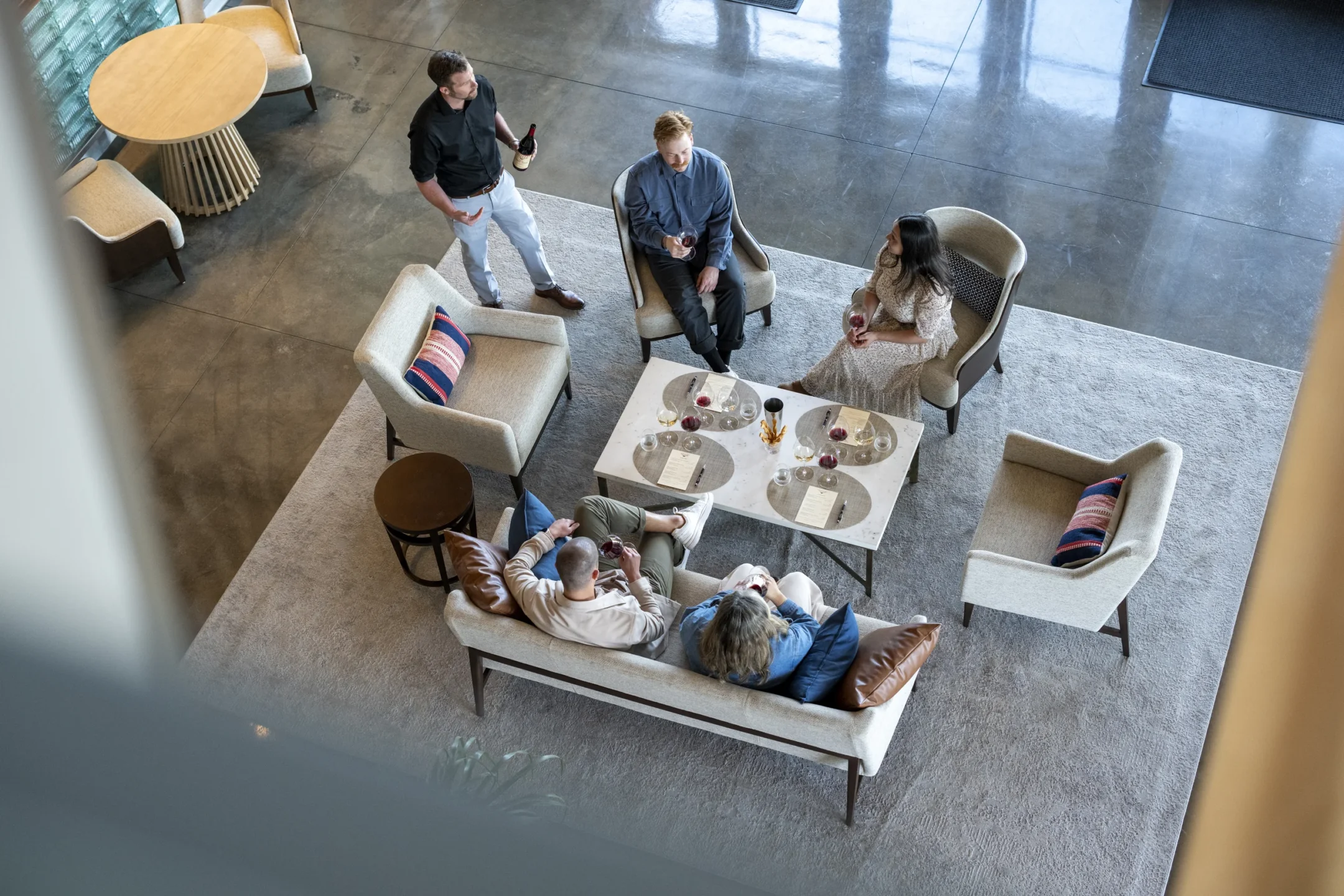 Five people are gathered in a modern lounge, sitting and standing around a coffee table with drinks, viewed from above.