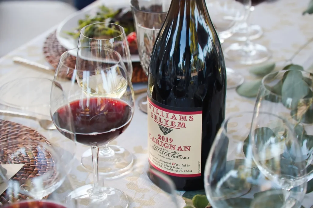 A bottle of 2019 Carignan red wine and two filled wine glasses sit on a set dining table with plates, utensils, and greenery.