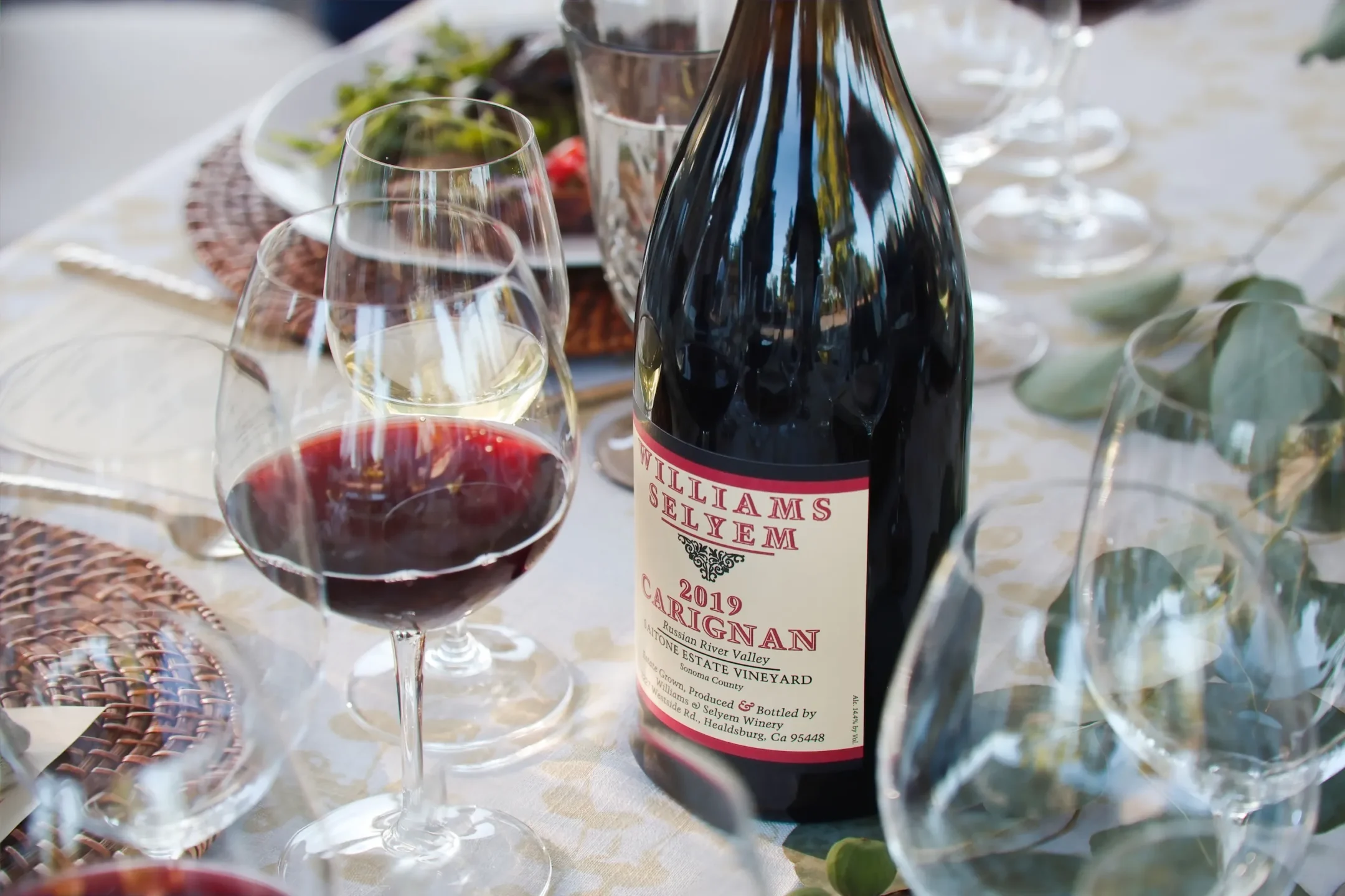 A bottle of 2019 Carignan red wine and two filled wine glasses sit on a set dining table with plates, utensils, and greenery.