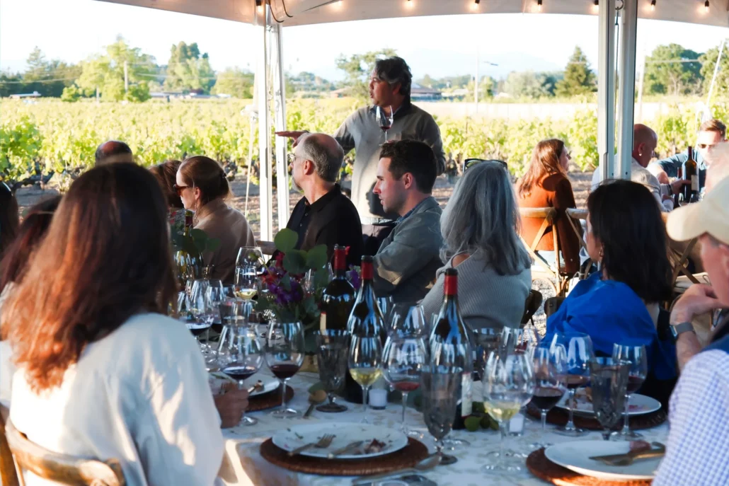 A group of people sit at a long table set with wine glasses and plates under a canopy, with a vineyard visible in the background.