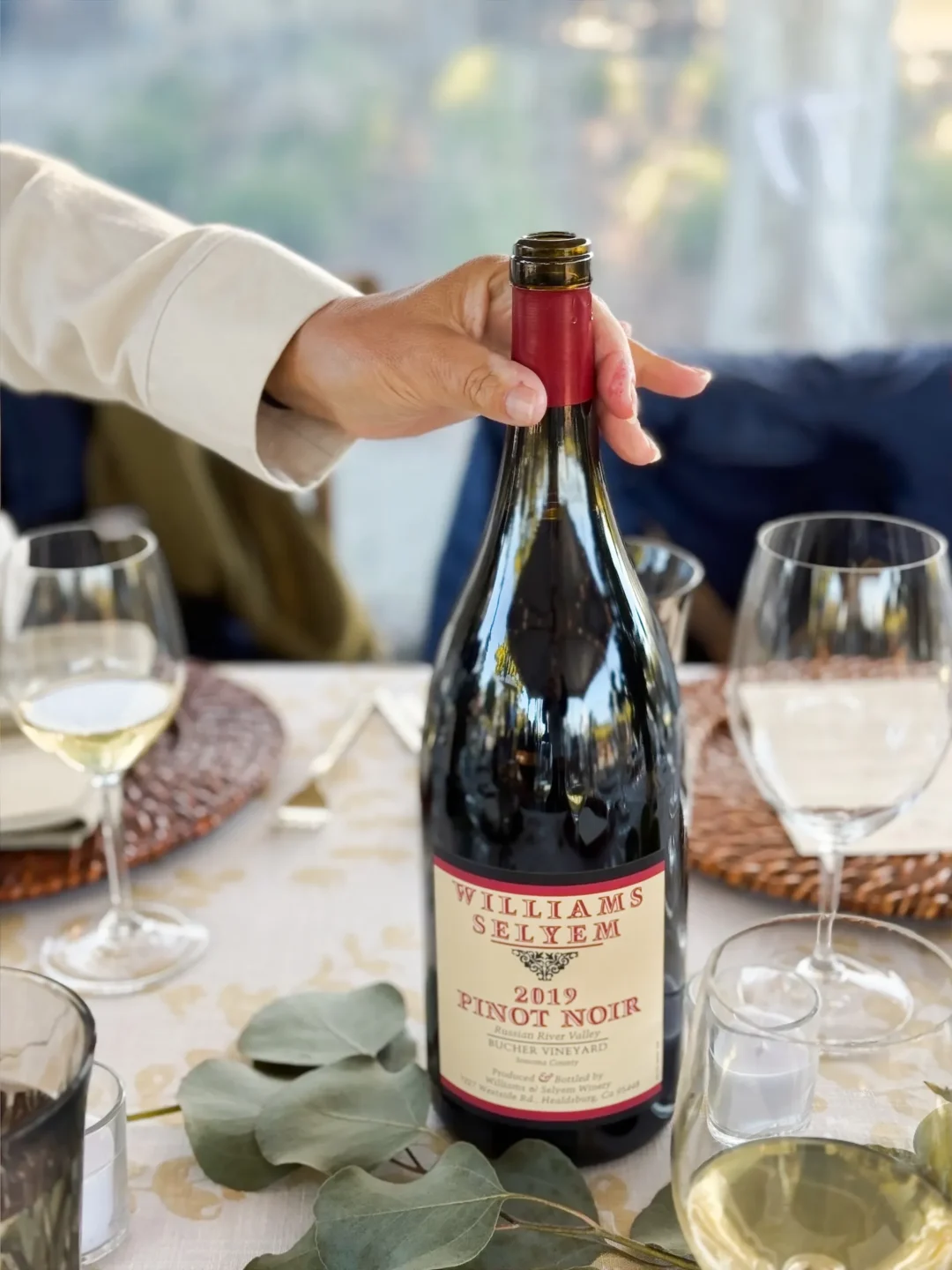 A hand holds a bottle of 2019 Williams Selyem Pinot Noir on a set dining table with wine glasses and greenery.