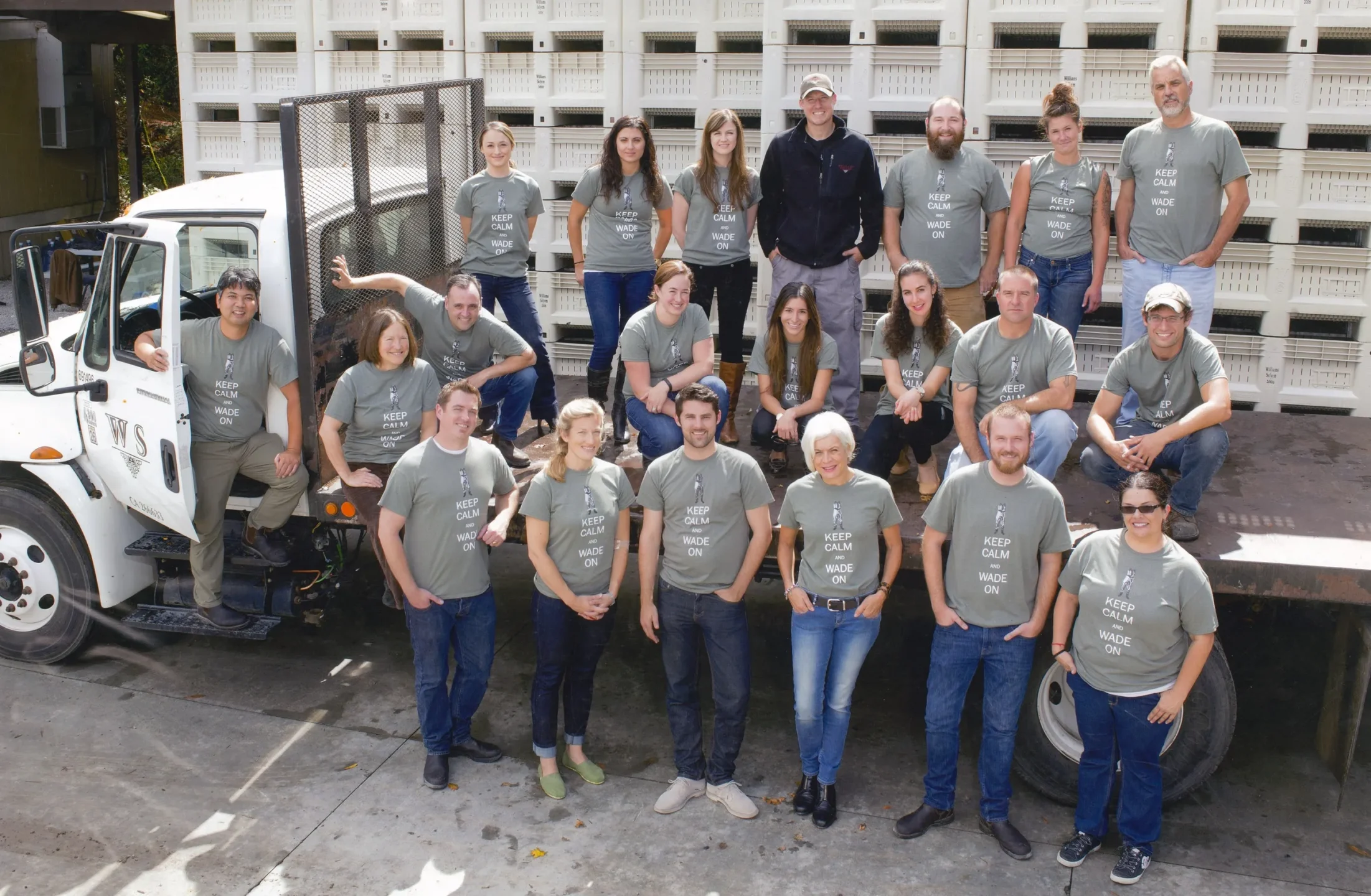A group of people wearing matching gray T-shirts poses together outdoors near a flatbed truck loaded with white crates.