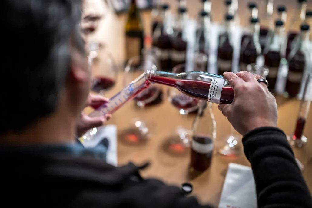 A person pours red liquid from a bottle into a graduated cylinder on a table with several bottles and glasses containing red liquid.