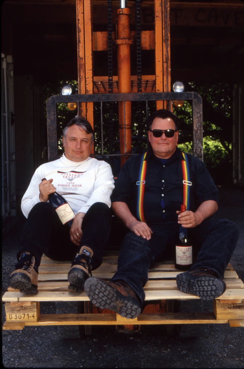 Williams Selyem Winery founders sit on a wooden pallet held by a forklift, each holding a bottle of Williams Selyem winemaking; one wears a white shirt, the other wears dark clothes with rainbow suspenders.