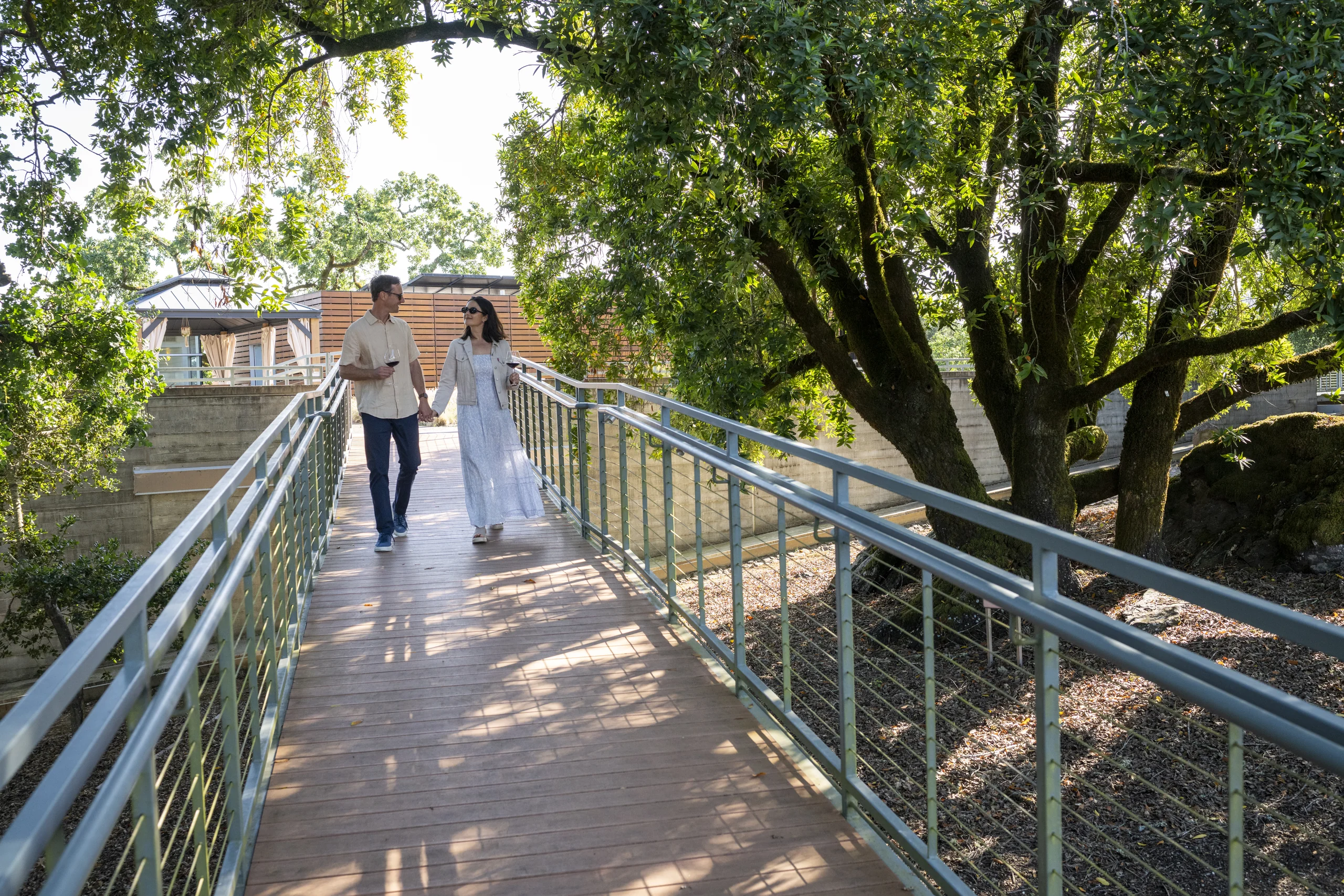 A man and woman hold hands at Williams Selyem Russian River Valley winery while walking on a wooden pedestrian bridge surrounded by trees and greenery on a sunny day.