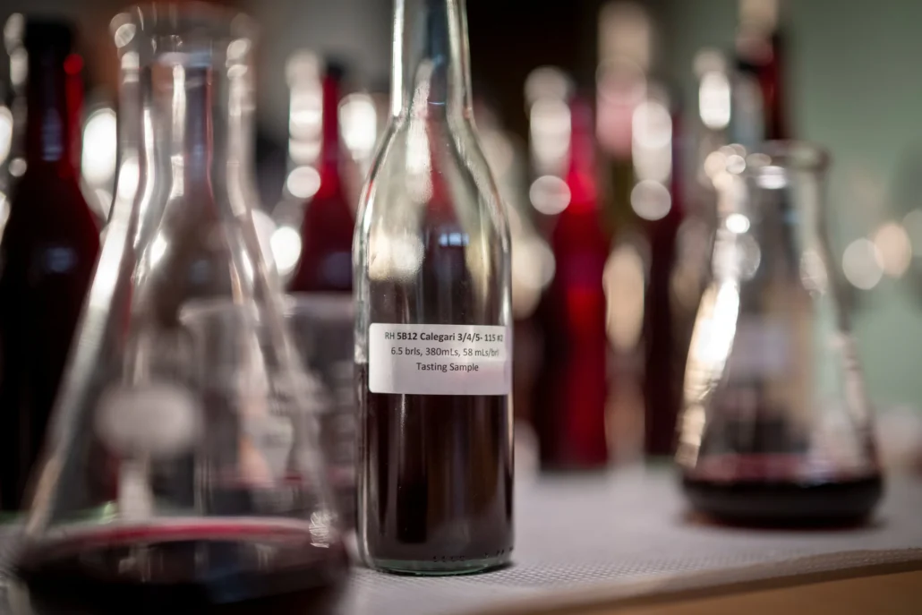 A clear glass bottle filled with dark liquid labeled "Tasting Sample" stands among other bottles and flasks, all containing red liquids, on a countertop.